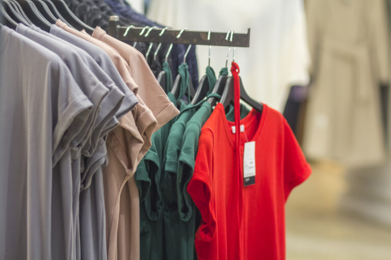 Row of colorful new modern unisex cotton t-shirts with price tags hanging on rack on blurred shop interior background. Fashion, sale and shopping concept
